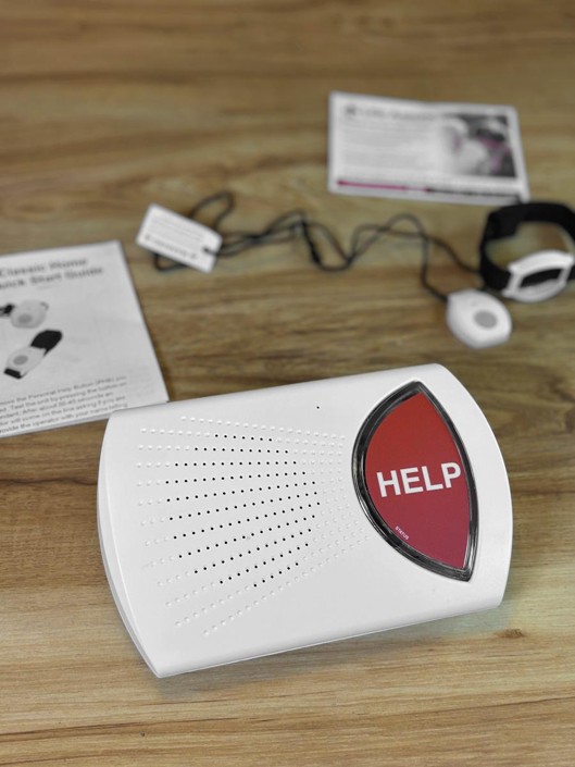 A white home base station with a large red HELP button on a wooden table, the printed quick start guide, a pendant on a black cord and a strap wristband set out behind it
